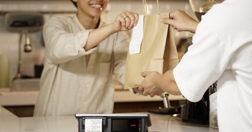 A smiling cashier handing a paper bag to a customer over the counter in a cozy cafe setting.