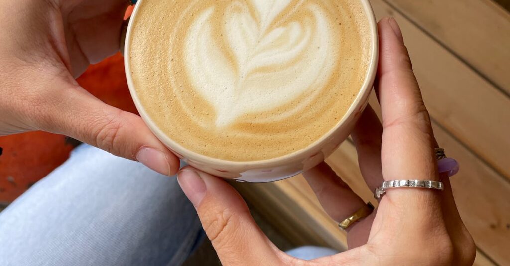 Top view of hands holding a cup of latte with elegant latte art on wooden seating.