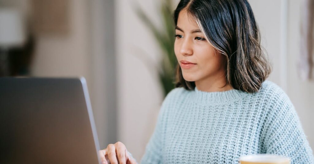 Confident young Latin American woman in casual clothes working remotely on netbook at table near mug with coffee in light apartment with plant with green leaves near wall